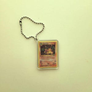 May include: A Charizard Pokémon trading card keychain. The card is encased in clear plastic and attached to a silver ball chain. The card features a Charizard illustration with text and a 100 HP rating.