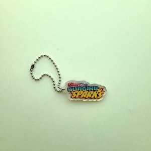 May include: A clear keychain with the text "Scarlet & Violet Surging Sparks" in blue, yellow, and red. The keychain is attached to a silver ball chain.