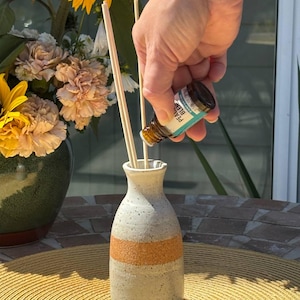May include: A ceramic essential oil diffuser with a tan band, filled with wooden reeds. A hand is pouring oil from a small bottle labeled "PEACE BLEND" into the diffuser. Sunflowers and other flowers are in a vase in the background.