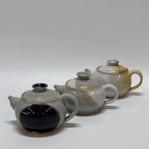 May include: Three ceramic teapots arranged in a diagonal line. The front teapot is light gray with a large, dark purple circular patch. The middle teapot features a light gray and white glaze with a tan streak. The back teapot is tan and white.
