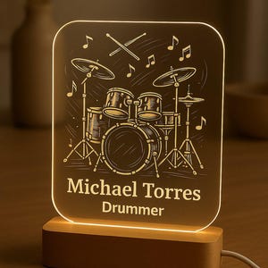 May include: An illuminated night light with a drum kit design and musical notes. The name "Michael Torres" and the word "Drummer" are displayed. The light is set on a wooden base, with a cable attached.