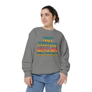 May include: A grey crewneck sweatshirt featuring the text "NOT EVERYONE GETS AN EXPLANATION" printed in a bold, multi-coloured gradient font that transitions from yellow to orange and pink, paired with blue denim jeans.
