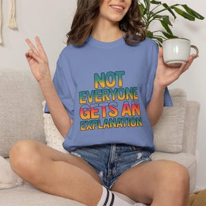 May include: A periwinkle blue short-sleeve t-shirt featuring the text NOT EVERYONE GETS AN EXPLANATION in a bold, multi-colored gradient font that transitions from yellow to orange to pink and teal, printed on the center of the chest.
