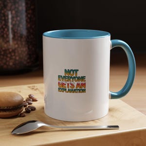 May include: A white ceramic mug with a blue interior and handle. The front of the mug features the text "NOT EVERYONE GETS AN EXPLANATION" printed in a retro-style, multi-colored gradient font with shades of yellow, orange, and green.