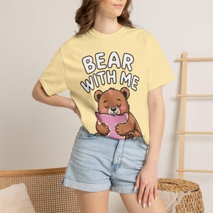 May include: A pale yellow short-sleeve t-shirt featuring a graphic of a brown bear holding a pink pillow. Above the bear, the text reads BEAR WITH ME in bold, black, rounded capital letters. The shirt has a crew neckline and a standard fit.