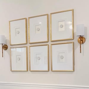 May include: Six framed artworks with gold trim and white mounts are displayed on a wall. Each frame features a white square with a circular design. Two sconces with white shades and gold fixtures are positioned near the artwork.