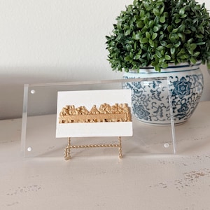 May include: A gold-colored relief sculpture of the Last Supper on a white background, displayed in a clear acrylic frame. The artwork is supported by a small, gold-toned metal stand. A potted plant in a blue and white patterned pot is in the background.