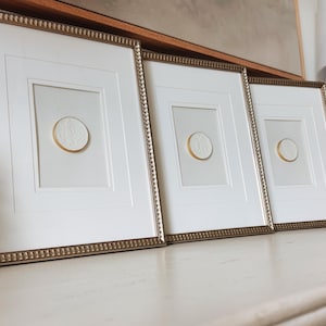 Può includere: Tre opere d'arte incorniciate con passe-partout bianchi e cornici dorate. Ogni cornice presenta un accento circolare testurizzato color oro al centro. Le cornici sono esposte su una superficie bianca, creando un set decorativo coerente.