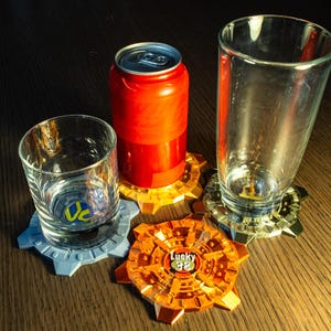 May include: A collection of drinkware and coasters. A red soda can and two clear glasses sit on gear-shaped coasters in gold, silver, and orange. A smaller glass sits on a gray coaster. The orange coaster has the text "Lucky 38".