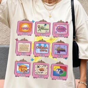 May include: Ivory-colored t-shirt featuring a grid of pink television screens, each displaying a different show title, including "Raven" and "High School Musical." The shirt has short sleeves and a relaxed fit. The text "Original Design by Lucky Lime Co" is at the bottom.