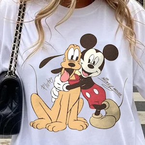 May include: White t-shirt with a cartoon illustration of Mickey Mouse embracing Pluto. Mickey Mouse is in red shorts, and Pluto is light brown. The words "Mickey Mouse" and "Pluto" are printed on the shirt.