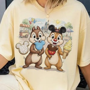 May include: A light yellow oversized t-shirt featuring a colorful graphic of Chip and Dale enjoying treats in front of a castle. Chip holds a blue ice cream, Dale holds a Mickey Mouse ice cream, and a donut is also visible.