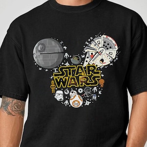 Pode incluir: Camiseta preta com um design de Star Wars em forma de orelhas do Mickey Mouse. O design inclui a Estrela da Morte, a Millennium Falcon e vários personagens de Star Wars. As palavras "STAR WARS" são em amarelo.