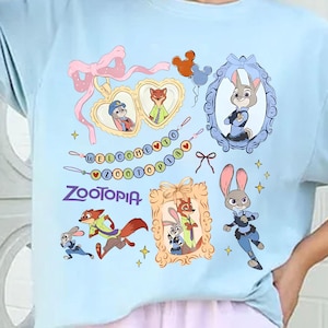May include: Light blue t-shirt featuring Zootopia characters and the word "Zootopia". The design includes framed images of Judy Hopps and Nick Wilde, along with other characters and decorative elements.