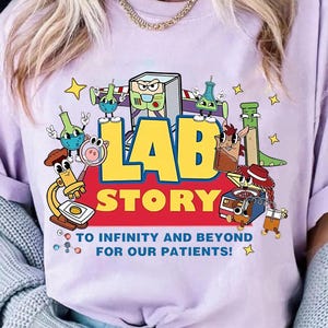 May include: A light purple t-shirt with a colorful graphic that reads "LAB STORY" with cartoon lab equipment and characters. The text "TO INFINITY AND BEYOND FOR OUR PATIENTS!" is also printed on the shirt.