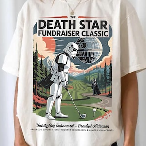 Może przedstawiać: Kremowa koszulka z grafiką Stormtroopera grającego w golfa, z Gwiazdą Śmierci w tle. Tekst brzmi "The Death Star Fundraiser Classic" i "Charity Golf Tournament - Beautiful Alderaan".