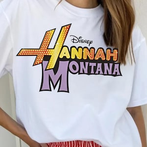 以下が含まれることがあります： 黄色、オレンジ、紫色のハンナ・モンタナのロゴが入った白いTシャツ。ディズニーのロゴが名前の上にあります。シャツは半袖です。