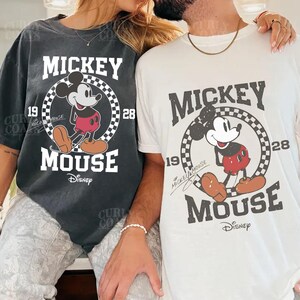 Peut inclure: Deux t-shirts avec un motif Mickey Mouse. L'un est gris fonc&eacute;, l'autre est blanc. Le motif comprend les mots "MICKEY MOUSE" et les ann&eacute;es "19" et "28", avec un cercle &agrave; carreaux autour de Mickey Mouse. Le logo Disney est en bas.
