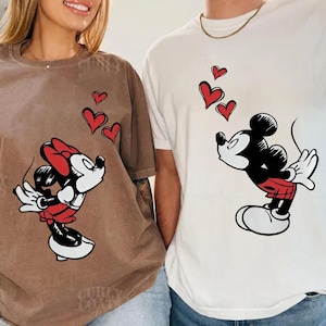May include: Two t-shirts with cartoon character designs. One is brown with Minnie Mouse, red hearts, and a red bow. The other is white with Mickey Mouse and red hearts. The shirts are a matching set.