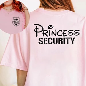 May include: A light pink t-shirt with the words "PRINCESS SECURITY" printed in black on the back. The front of the shirt features a small emblem with the words "PRINCESS SECURITY" and a crown design.
