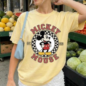 May include: A pale yellow t-shirt with a Mickey Mouse graphic. The design features the words "Mickey Mouse" in red, with a black and white chequered circle around Mickey. The text "EST. 1928" is also included.