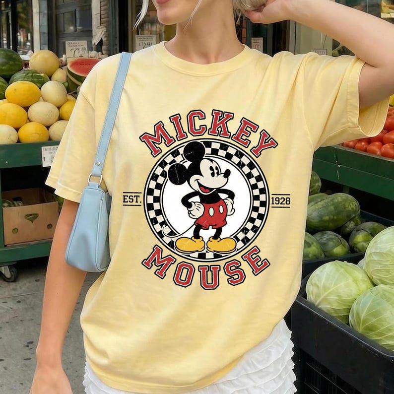 May include: A pale yellow t-shirt with a Mickey Mouse graphic. The design features the words "Mickey Mouse" in red, with a black and white chequered circle around Mickey. The text "EST. 1928" is also included.