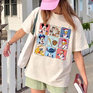 May include: Cream-colored t-shirt featuring a grid of cartoon characters in baseball uniforms. Mickey Mouse, Minnie Mouse, Chip and Dale, Donald Duck, Goofy, and Stitch are among the characters. A baseball, bat, and glove are also featured.