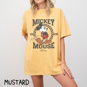 Peut inclure: T-shirt jaune moutarde avec un motif Mickey Mouse vieilli. Le design comprend les mots "Mickey Mouse" et les ann&eacute;es "19" et "28". Le logo Disney est en bas du motif.