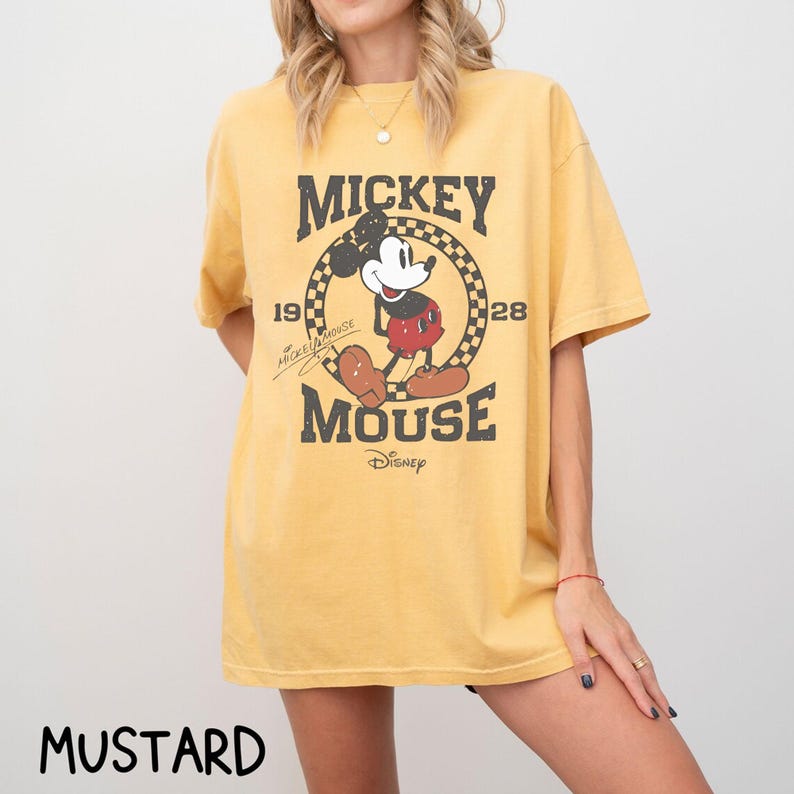 Peut inclure: T-shirt jaune moutarde avec un motif Mickey Mouse vieilli. Le design comprend les mots "Mickey Mouse" et les ann&eacute;es "19" et "28". Le logo Disney est en bas du motif.