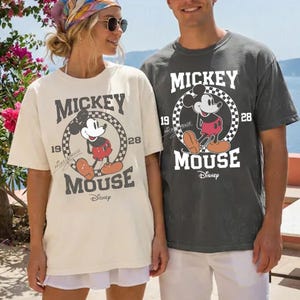 Peut inclure: Deux t-shirts avec un motif Mickey Mouse. L'un est de couleur cr&egrave;me, l'autre gris anthracite. Le motif comprend Mickey Mouse, les mots "Mickey Mouse" et le logo Disney. Le motif est entour&eacute; d'un motif &agrave; carreaux et des chiffres "19" et "28".