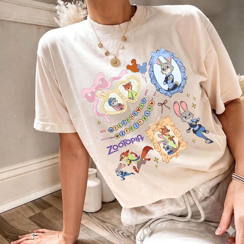 May include: Cream-colored t-shirt featuring colorful cartoon characters and the word "Zootopia". The design includes framed portraits of characters, a pink bow, and the phrase "Welcome to Zootopia" in a playful font.