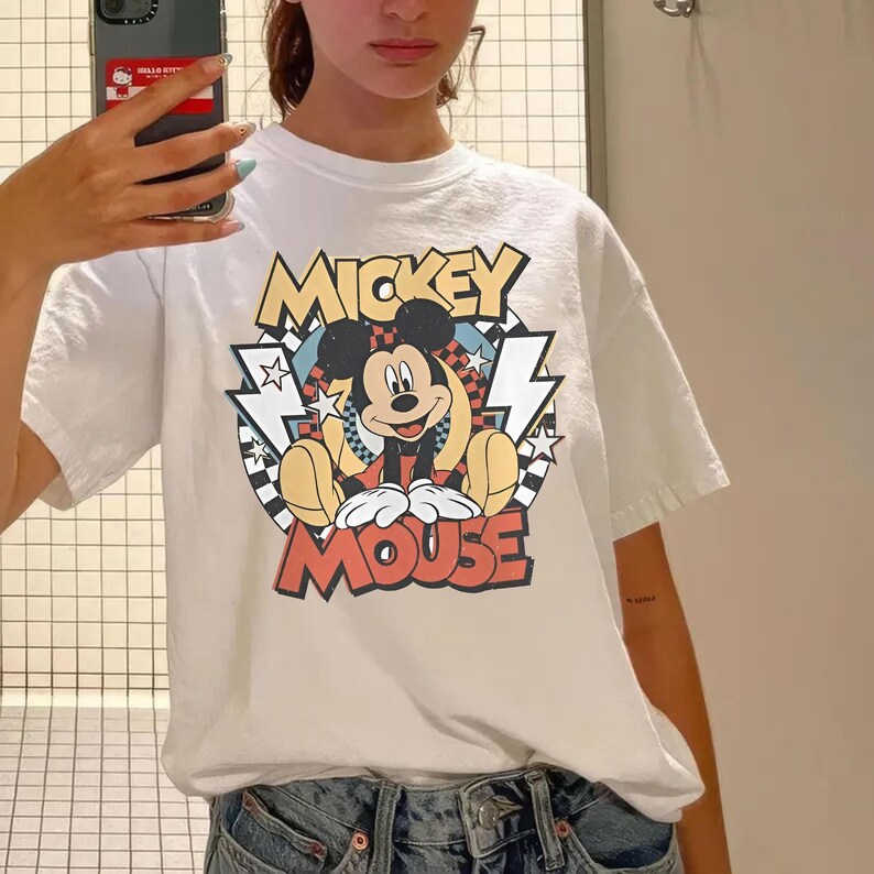 May include: White t-shirt featuring a graphic of Mickey Mouse. The design includes the words "Mickey Mouse" in yellow and red, with Mickey in the center. The shirt has short sleeves and a crew neck.