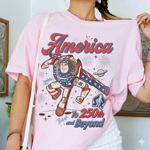 May include: A light pink t-shirt featuring a graphic of a space ranger character in a red, white, and blue suit. The text reads America To 250th and Beyond, accompanied by illustrations of fireworks, a planet, and a rocket ship emblem in blue and red.