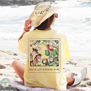 K&ouml;nnte beinhalten: Hellgelbes T-Shirt mit einem rechteckigen Aufdruck mit Figuren aus dem Film Toy Story. Der Aufdruck zeigt Woody, Buzz Lightyear und andere Charaktere. Der Text "YOU'VE GOT A FRIEND IN ME" steht darunter. Das T-Shirt wird von einer Person am Strand getragen.