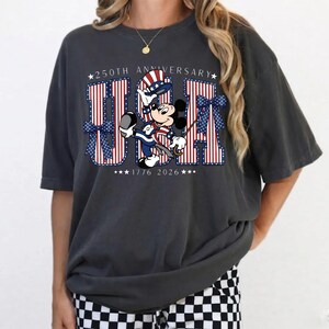 May include: Dark grey t-shirt with the word "USA" in red and white stripes, blue bows, and stars. Mickey Mouse is depicted in patriotic clothing. The shirt celebrates the 250th anniversary, with the years 1776 and 2026.