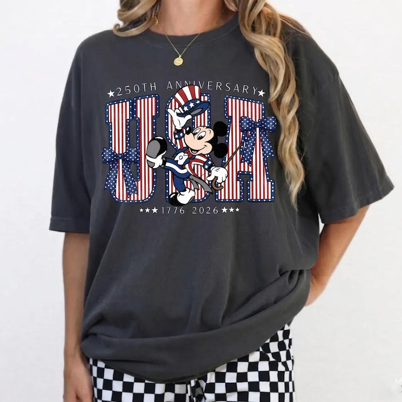 May include: Dark grey t-shirt with the word "USA" in red and white stripes, blue bows, and stars. Mickey Mouse is depicted in patriotic clothing. The shirt celebrates the 250th anniversary, with the years 1776 and 2026.