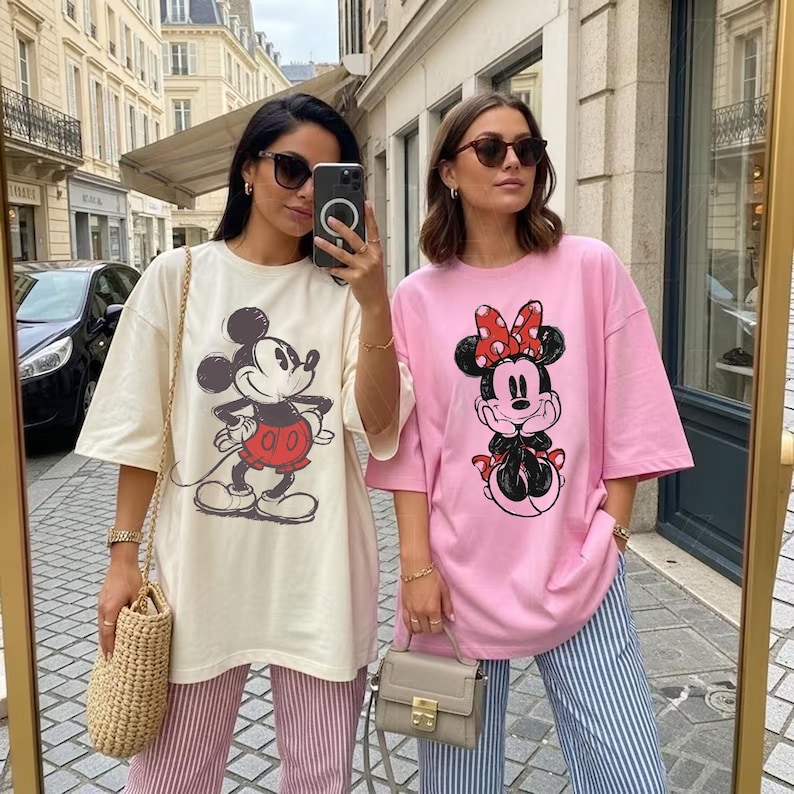 May include: Two women wearing oversized t-shirts with Disney character graphics. One shirt is cream-colored with Mickey Mouse, the other is pink with Minnie Mouse. Both are paired with striped pants. The women are in an outdoor setting.