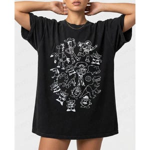 May include: Black t-shirt featuring a white cartoon illustration of characters from a popular animated movie. The design includes a cowboy, space ranger, and other recognizable figures. The shirt has a relaxed fit.