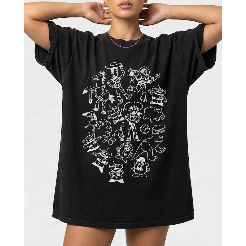 May include: Black t-shirt featuring a white cartoon illustration of characters from a popular animated movie. The design includes a cowboy, space ranger, and other recognizable figures. The shirt has a relaxed fit.