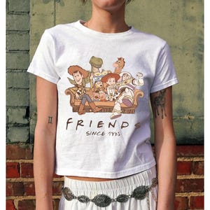 May include: White t-shirt featuring a graphic of characters from the animated film Toy Story, seated on a couch. The text "FRIENDS SINCE 1995" is printed below the image. The shirt is short-sleeved.