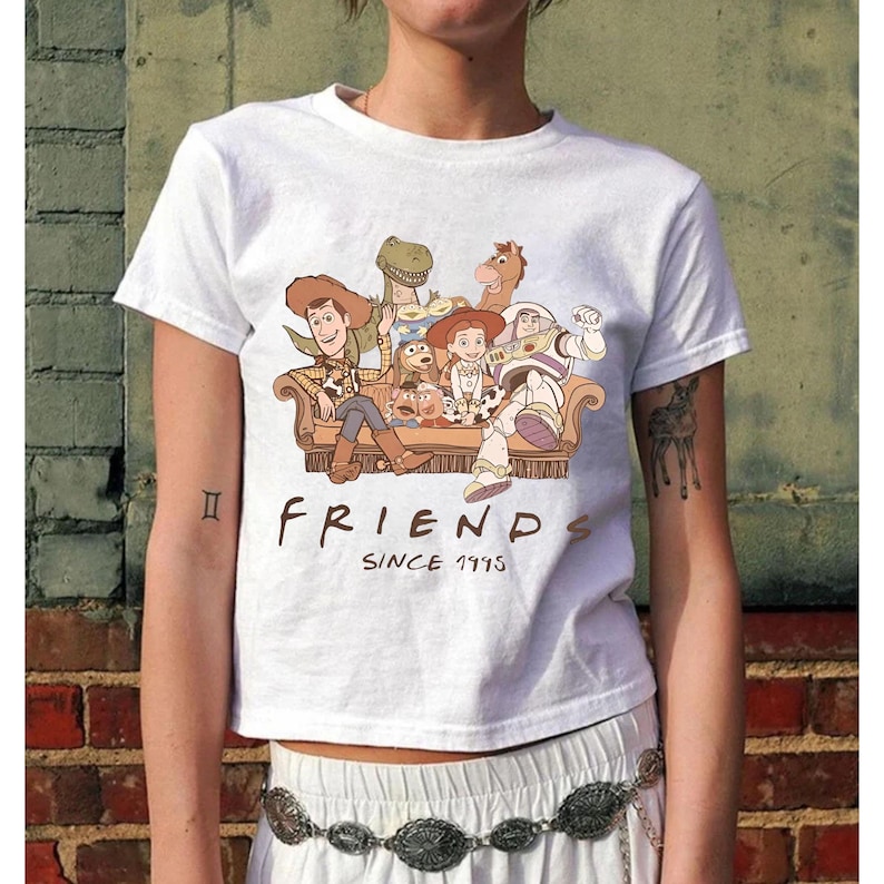 May include: White t-shirt featuring a graphic of characters from the animated film Toy Story, seated on a couch. The text "FRIENDS SINCE 1995" is printed below the image. The shirt is short-sleeved.