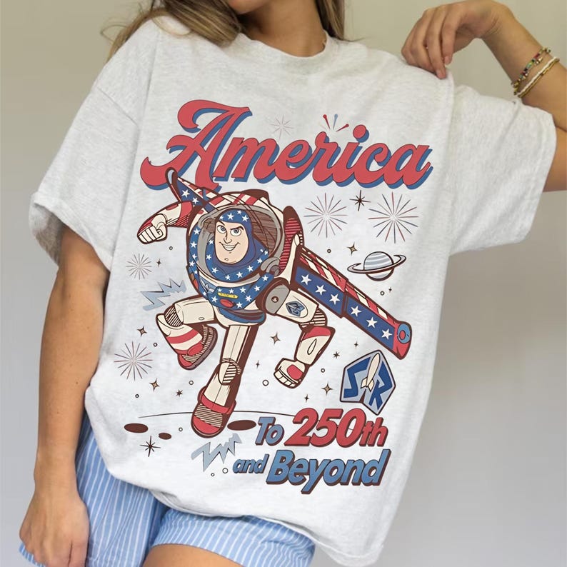 May include: A light gray short-sleeve t-shirt featuring a graphic of a space ranger character in a red, white, and blue suit. The text reads America at the top and To 250th and Beyond at the bottom, surrounded by star and firework illustrations.