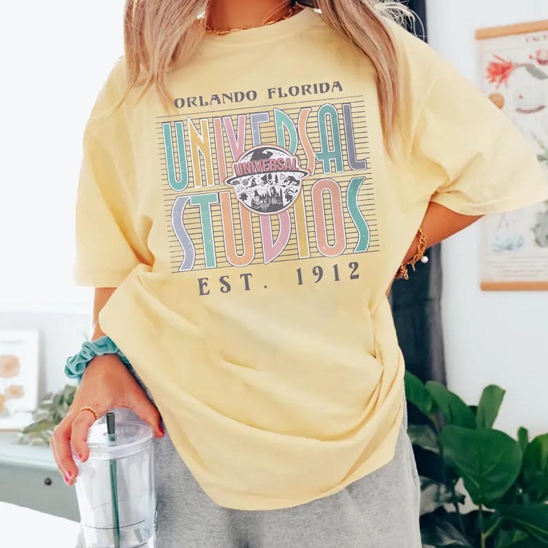 May include: A light yellow t-shirt with the text "ORLANDO FLORIDA UNIVERSAL STUDIOS EST. 1912" in a colorful, retro design. The shirt has short sleeves and a relaxed fit, perfect for a casual day out.