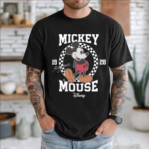 Peut inclure: T-shirt noir avec un dessin de Mickey Mouse. Le design comprend les mots "Mickey Mouse" en blanc, un cercle &agrave; carreaux et les ann&eacute;es "19" et "28". Le logo Disney est en bas.