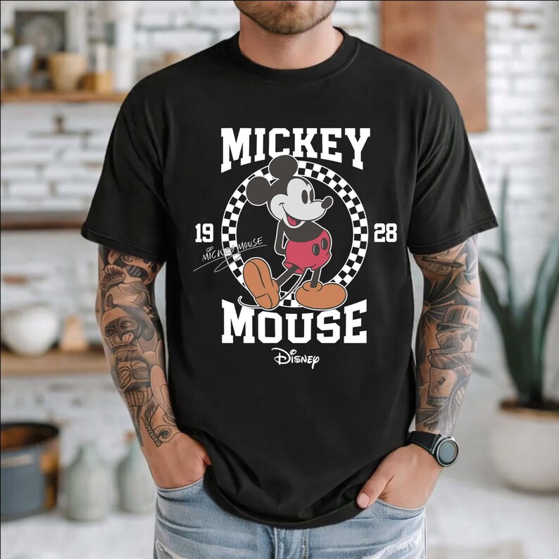 Peut inclure: T-shirt noir avec un dessin de Mickey Mouse. Le design comprend les mots "Mickey Mouse" en blanc, un cercle &agrave; carreaux et les ann&eacute;es "19" et "28". Le logo Disney est en bas.