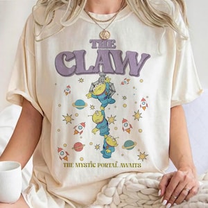 Puede incluir: Una camiseta de color crema con un estampado gráfico que incluye el texto THE CLAW en la parte superior y THE MYSTIC PORTAL AWAITS en la parte inferior. El diseño muestra tres personajes alienígenas verdes, cohetes, planetas y estrellas.