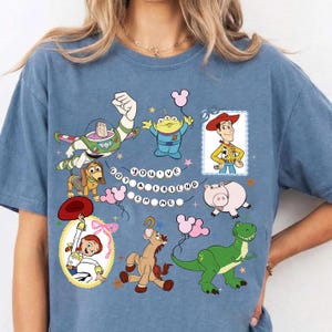 May include: A blue t-shirt featuring a graphic print of Toy Story characters including Buzz Lightyear, Woody, Jessie, Slinky Dog, Rex, Hamm, and an Alien. The text "YOU'VE GOT A FRIEND IN ME" is displayed in the center with small star and balloon accents.