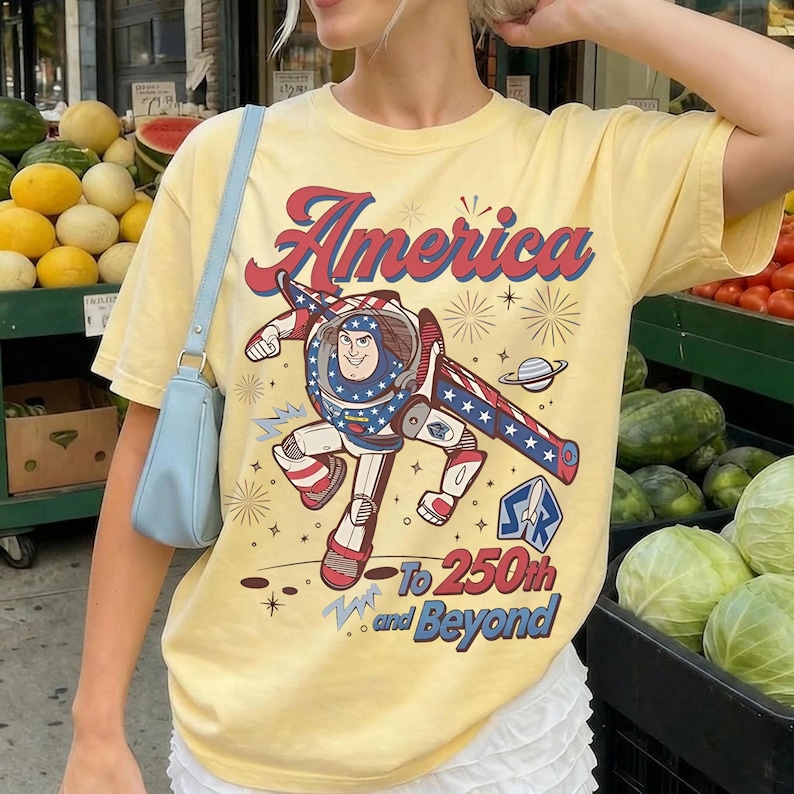 May include: A pale yellow short-sleeve t-shirt featuring a graphic of the character Buzz Lightyear with red, white, and blue patriotic accents. The text reads America To 250th and Beyond, accompanied by illustrations of fireworks and a planet.