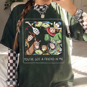 K&ouml;nnte beinhalten: Dunkelgr&uuml;nes T-Shirt mit einem rechteckigen Aufdruck von Toy Story-Charakteren, darunter Woody und Buzz Lightyear. Der Text "YOU'VE GOT A FRIEND IN ME" ist darunter gedruckt.