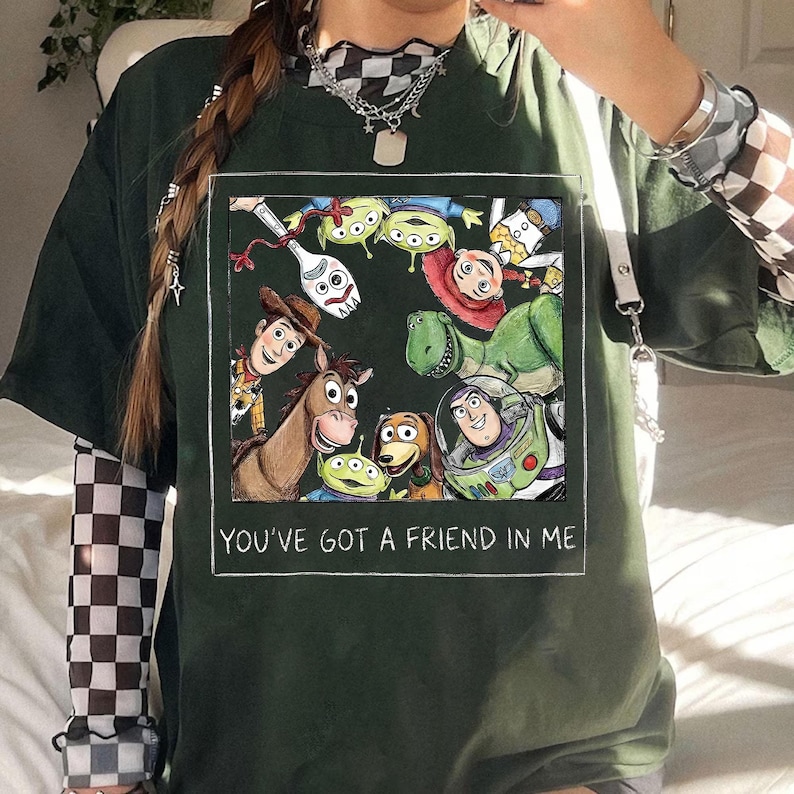 K&ouml;nnte beinhalten: Dunkelgr&uuml;nes T-Shirt mit einem rechteckigen Aufdruck von Toy Story-Charakteren, darunter Woody und Buzz Lightyear. Der Text "YOU'VE GOT A FRIEND IN ME" ist darunter gedruckt.